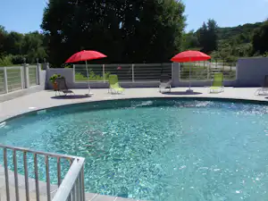 Camping-Pool mit türkisfarbenem Wasser, roten Sonnenschirmen und Liegestühlen auf der Terrasse hinter einem weißen Zaun.