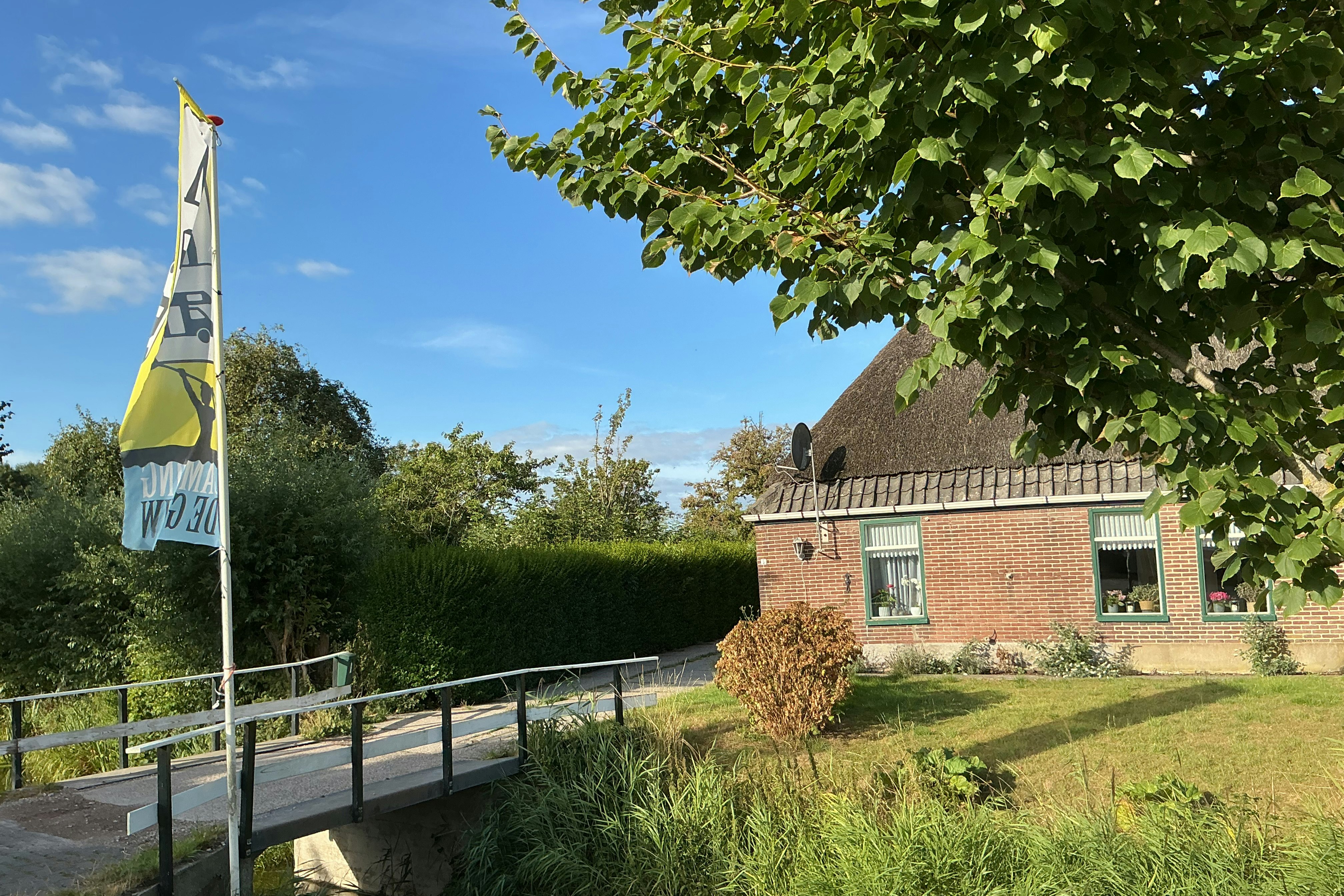 Gelbes Banner am Mast neben einem Fußweg, dahinter ein Backsteinhaus mit Terrasse und grüner Wiese unter blauem Himmel.