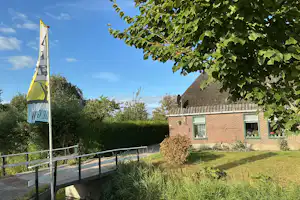 Gelbes Banner am Mast neben einem Fußweg, dahinter ein Backsteinhaus mit Terrasse und grüner Wiese unter blauem Himmel.