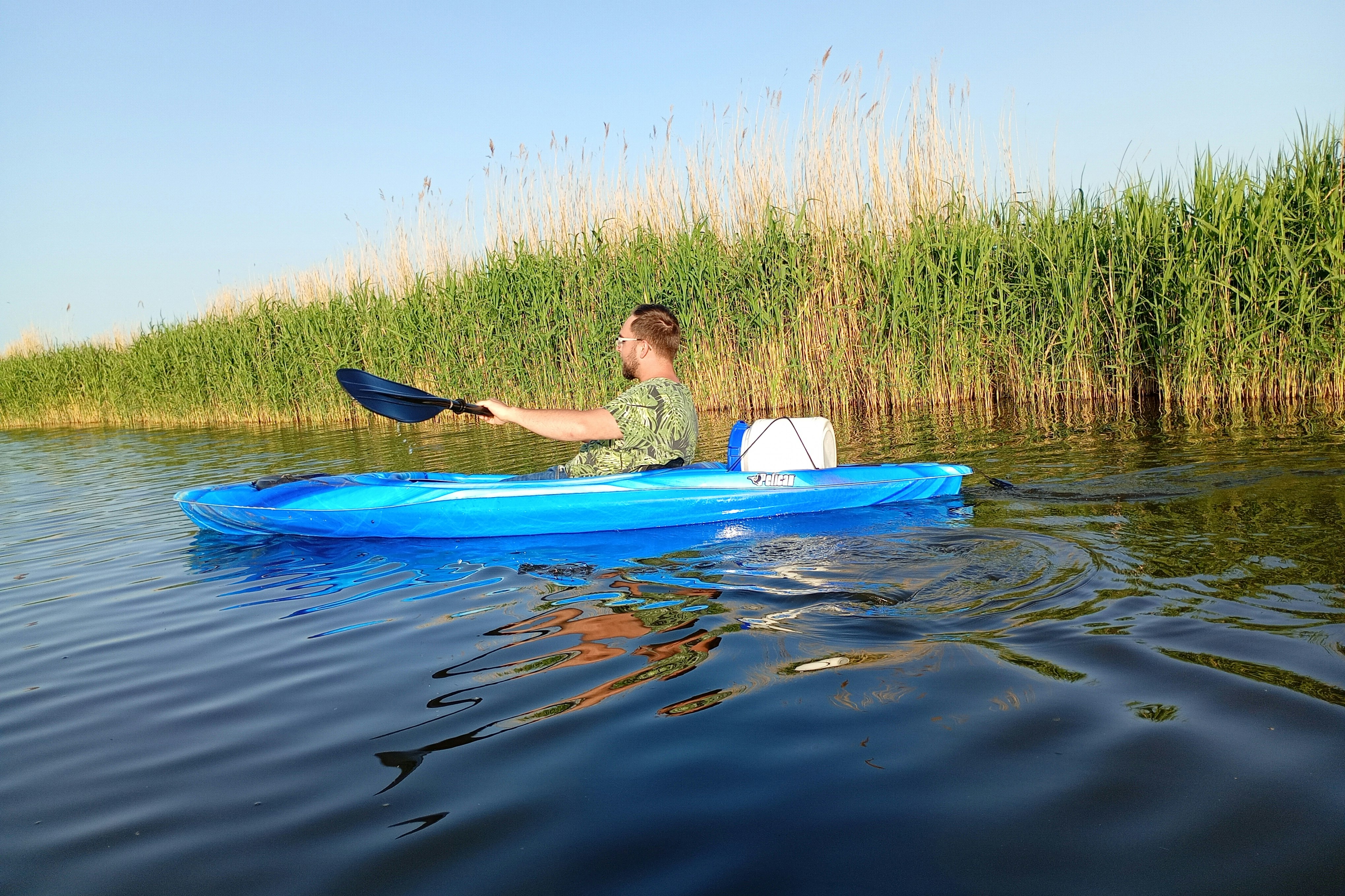 Eine Person paddelt in einem blauen Schlauchboot auf dem See, Schilfufer im Hintergrund.