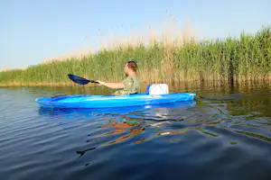 Eine Person paddelt in einem blauen Schlauchboot auf dem See, Schilfufer im Hintergrund.
