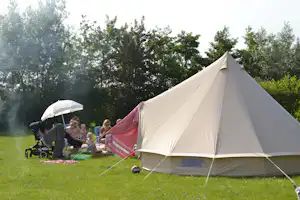Großes Campingzelt auf einer grünen Wiese, daneben ein Kinderwagen und ein Sonnenschirm mit sitzenden Personen.