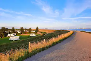 Campingplatz mit Wohnwagen auf der Wiese, Feldweg am Ufer und Segelboote in der Ferne.