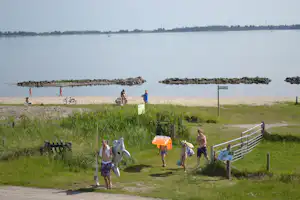 Kinder spielen auf dem Weg am Seeufer, im Hintergrund Wasser und Stege, daneben grünes Gras und Absperrgeländer.