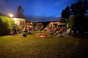 Campingplatz am Abend: Zelte, Wohnwagen und ein Lagerfeuer auf dem Rasen, umgeben von Bäumen und Laternen.