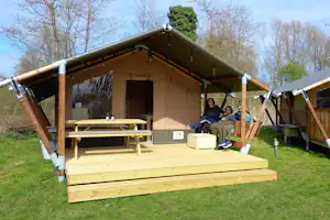 Glamping-Zelt mit Holzterrasse, Sitzbank und Tisch unter grünem Dach, im Hintergrund Bäume und Rasen.