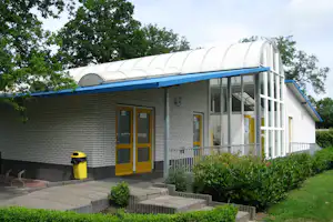 Campinggebäude mit Ziegelwand, gelben Türen und Glasfront, davor Gehweg und Geländer, im Hintergrund Bäume.