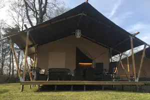 Glamping-Zelt mit Holzrahmen und Markise, davor Stühle auf einer Terrasse, im Hintergrund Bäume.