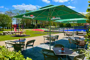 Terrasse mit Tischen und Stühlen unter grünem Sonnenschirm, daneben Spielplatz und Blick auf den Poolbereich.