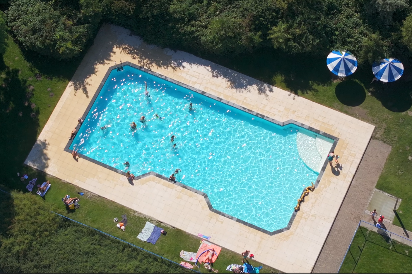 Großes Camping-Schwimmbecken mit blauem Wasser, Liegen und Sonnenschirmen auf einer Terrasse, umgeben von Bäumen und Rasen.