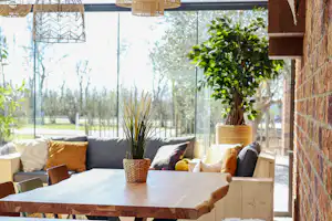 Überdachte Terrasse mit Holztisch und Sitzkissen, hängenden Lampen und Blick ins Grüne durch große Glasfronten.