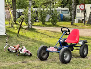 Spielzeug-Kindertraktor auf dem Rasen eines Campingplatzes, im Hintergrund Wohnwagen und Zelte zwischen Bäumen.