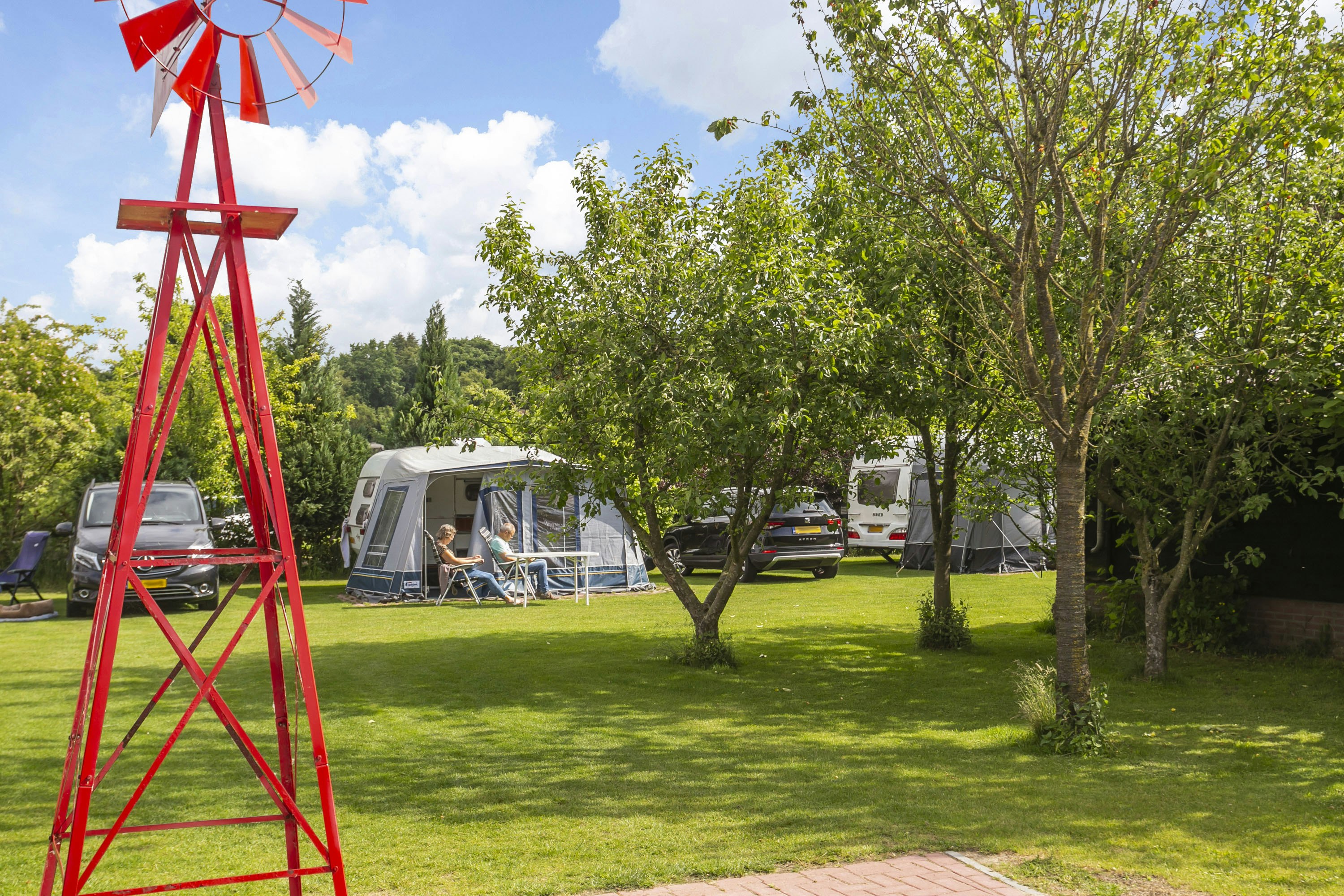 Roter Windmühlen-Deko auf dem Campingplatz, im Hintergrund Wohnwagen und Sitzgelegenheiten auf dem Rasen unter Bäumen.