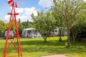Roter Windmühlen-Deko auf dem Campingplatz, im Hintergrund Wohnwagen und Sitzgelegenheiten auf dem Rasen unter Bäumen.