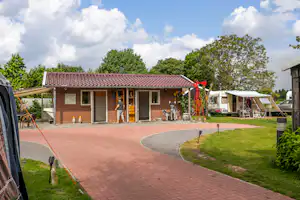 Reihenbungalow mit Terrasse und Sitzbereich, gepflasterter Weg und Rasenfläche, daneben Wohnwagen und Sonnenschirm im Campingbereich.