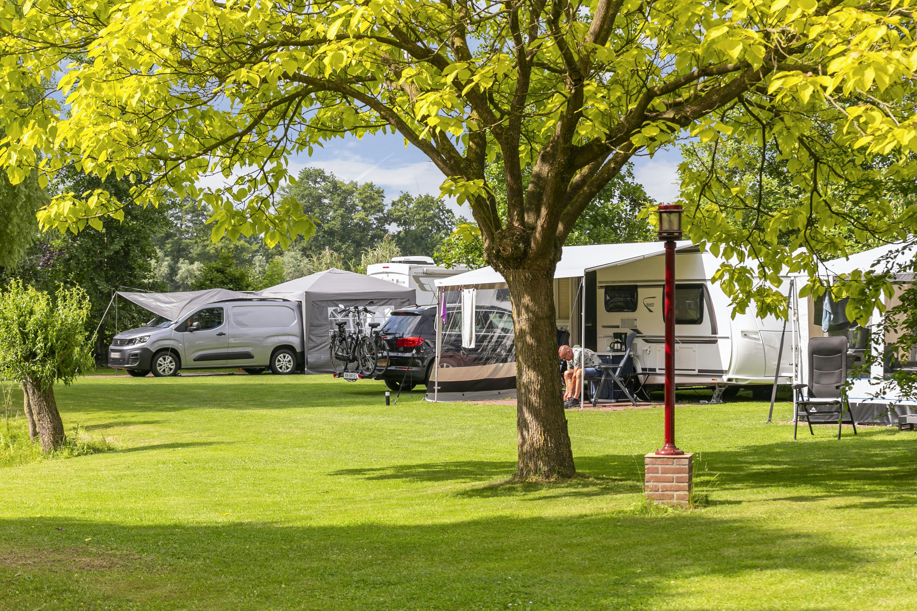 Wohnwagen und Wohnmobil auf einem Campingplatz, mit Campingstuhl und Tisch am Stellplatz, umgeben von Rasen und Bäumen.