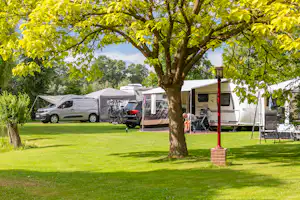 Wohnwagen und Wohnmobil auf einem Campingplatz, mit Campingstuhl und Tisch am Stellplatz, umgeben von Rasen und Bäumen.