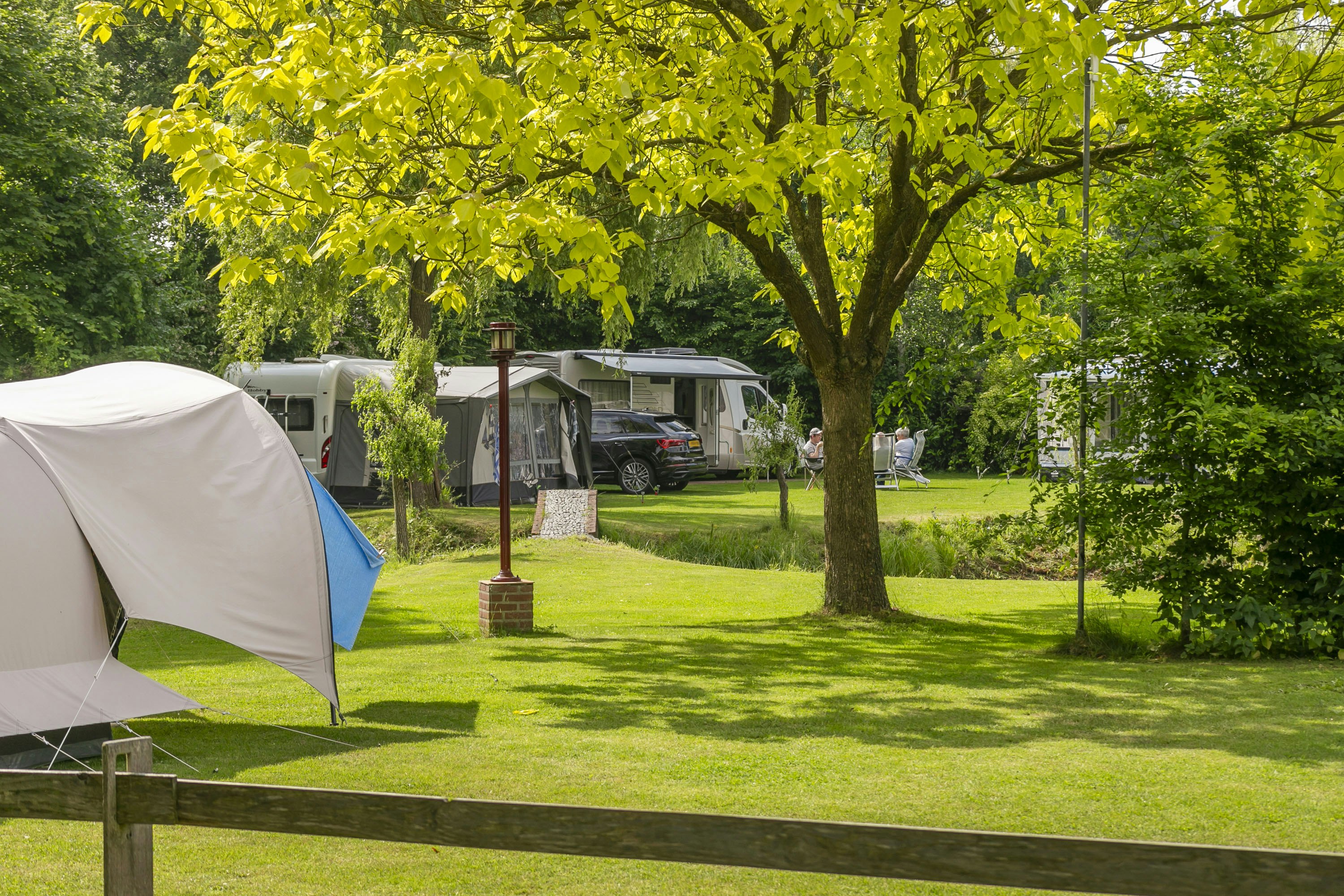 Zelt auf einem Campingplatz mit gepflegtem Rasen, daneben Wohnwagen und ein Auto, im Hintergrund Bäume und Hecken.