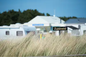 Campingplatz mit Wohnwagen und Zelten, im Vordergrund hohes Gras, im Hintergrund Gebäude und das Schild „Zuidveld“.