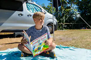 Camping De Kiekduun - Kind sitzt vor einem Standplatz und liest eine Zeitschrift