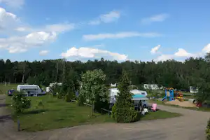 Campingplatz mit Wohnmobilen, Vorzelten und Sitzbereich, umgeben von Bäumen und einem Spielplatz mit Rutsche.