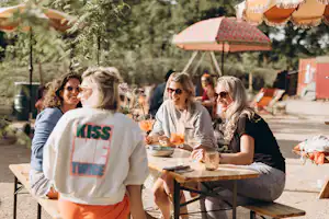 Gruppe am Camping-Tisch im Freien mit Getränken, Sonnenschirm und Zelten im Hintergrund.