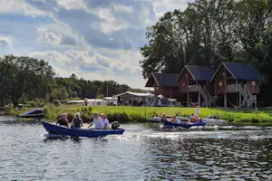 Boot mit Personen auf dem See vor einem Campingplatz mit Zelten, Rasen und Holzgebäuden am Ufer.