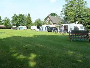 Wohnwagen und Zelte auf grünem Campingplatz, mit Fahrrädern und Sitzgelegenheiten vor einem Haus im Hintergrund.