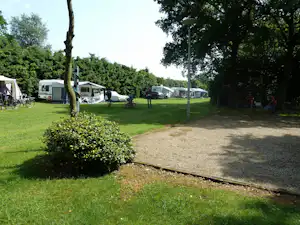 Campingplatz mit Wohnwagen und Zelten auf dem Rasen, Stühle und Fahrräder neben dem Weg, umgeben von Bäumen.