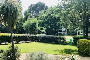 Grüne Parkanlage mit Bäumen, Hecken und Rasenfläche; im Hintergrund ein Spielbereich mit Toren und Absperrungen.