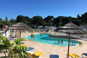 Camping-Pool mit Sonnenschirmen aus Stroh, Liegen und mehreren Personen im Wasser vor Bungalows und Bäumen.