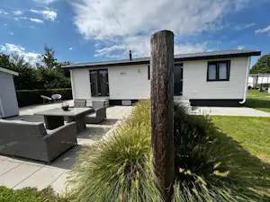 Weiße Mobilheim mit Terrasse, Sitzgruppe und gepflastertem Weg, davor Rasen und Ziergras im Garten.