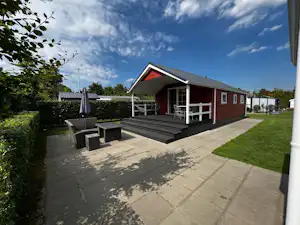 Rotes Campinghaus mit überdachter Terrasse, Sitzstufen und Gartenweg, daneben Hecken und Rasenfläche.