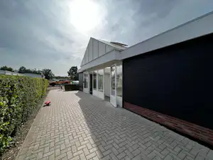 Haus am Campingplatz mit gepflasterter Terrasse, großen Fenstern und Hecke, im Hintergrund Stellplätze und Bäume.