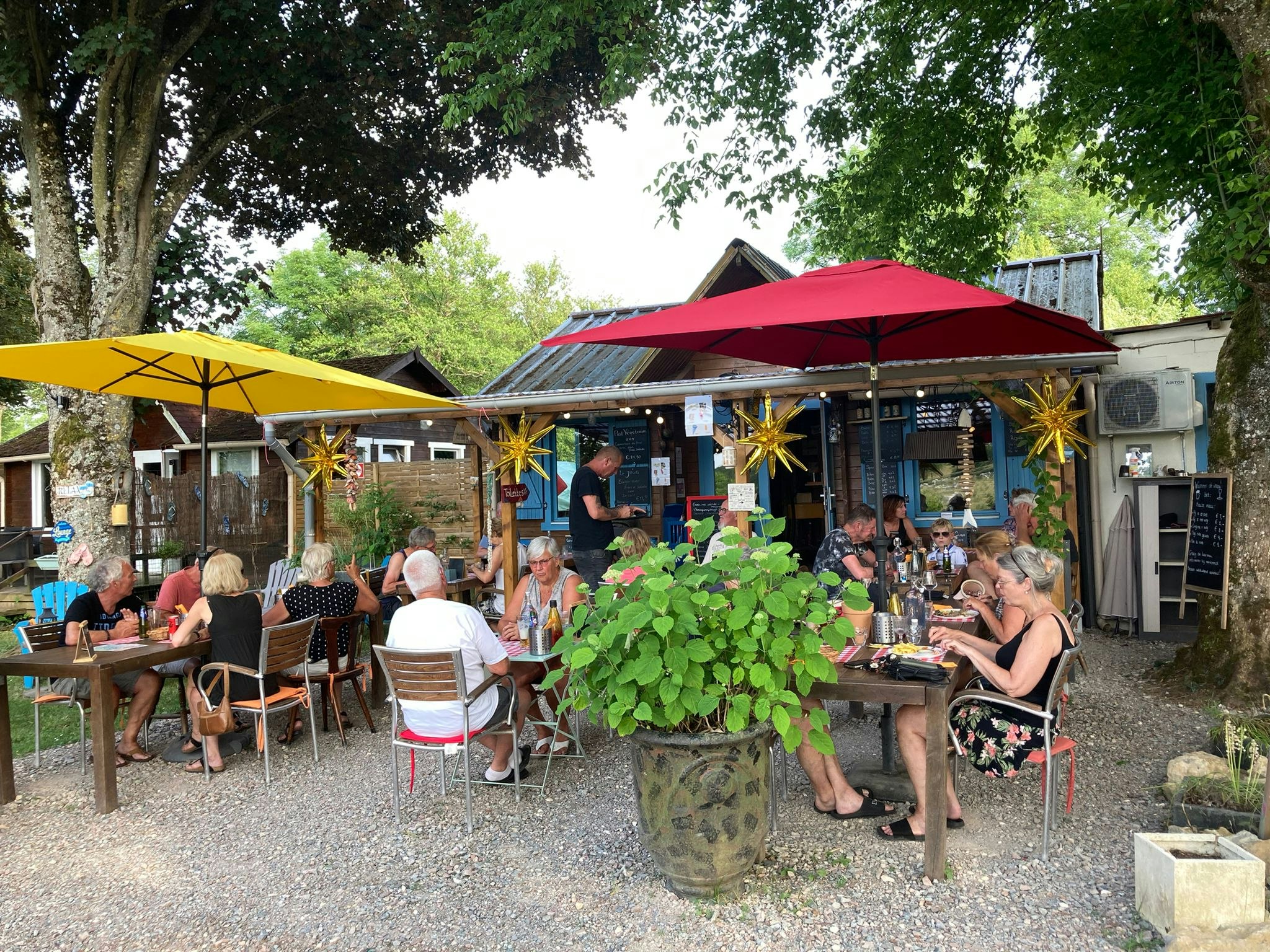 Terrasse am Campingplatz mit Gartenmöbeln, Sonnenschirmen und mehreren Personen vor einem Gebäude im Grünen.
