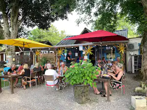 Terrasse am Campingplatz mit Gartenmöbeln, Sonnenschirmen und mehreren Personen vor einem Gebäude im Grünen.