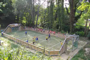 Volleyballfeld auf dem Campingplatz mit Holzumrandung, Netz und Spielern im Grünen zwischen hohen Bäumen.