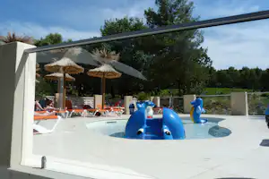 Kinderplanschbecken mit blauen Delfinfiguren am Pool, Liegen und Sonnenschirme auf der Terrasse, Bäume im Hintergrund.