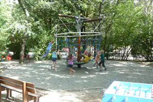 Kinder spielen auf einem Klettergerüst mit Seilen und Netz im Schatten der Bäume auf einem Kiesplatz.