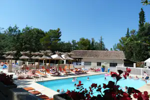 Poolbereich im Campingurlaub: Sonnenliegen, Strohdächer und Sonnenschirme am Becken, dahinter Bungalows und Bäume.