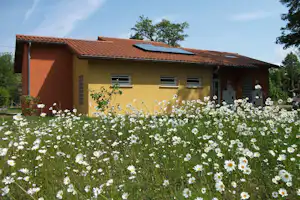 Gelbes Campinghaus mit rotem Ziegeldach und Solarpanel, davor ein Blumenbeet mit weißen Margeriten und grünen Büschen.