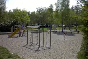 Spielplatz am Campingplatz mit Klettergerüst, Rutsche und Sandboden, umgeben von Bäumen und Wegen.