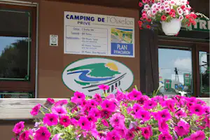 Blumenbeet mit pinken Petunien vor einem Camping-Infoboard „Camping de l’OiseIon“ am Eingang, daneben Fenster und Holzrahmen.