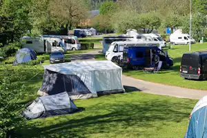 Zelte und Wohnwagen auf einem Campingplatz neben einem Weg, umgeben von Rasen, Bäumen und Bergen im Hintergrund.