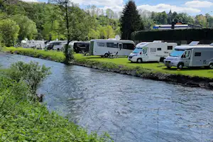 Wohnmobile und Campingbusse stehen am Flussufer auf dem Campingplatz, mit Bäumen und Rasenfläche im Hintergrund.
