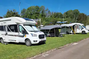 Wohnmobile und Zelte auf einem Campingplatz, mit Vorzelten, Campingstühlen und Rasenfläche neben der Straße.