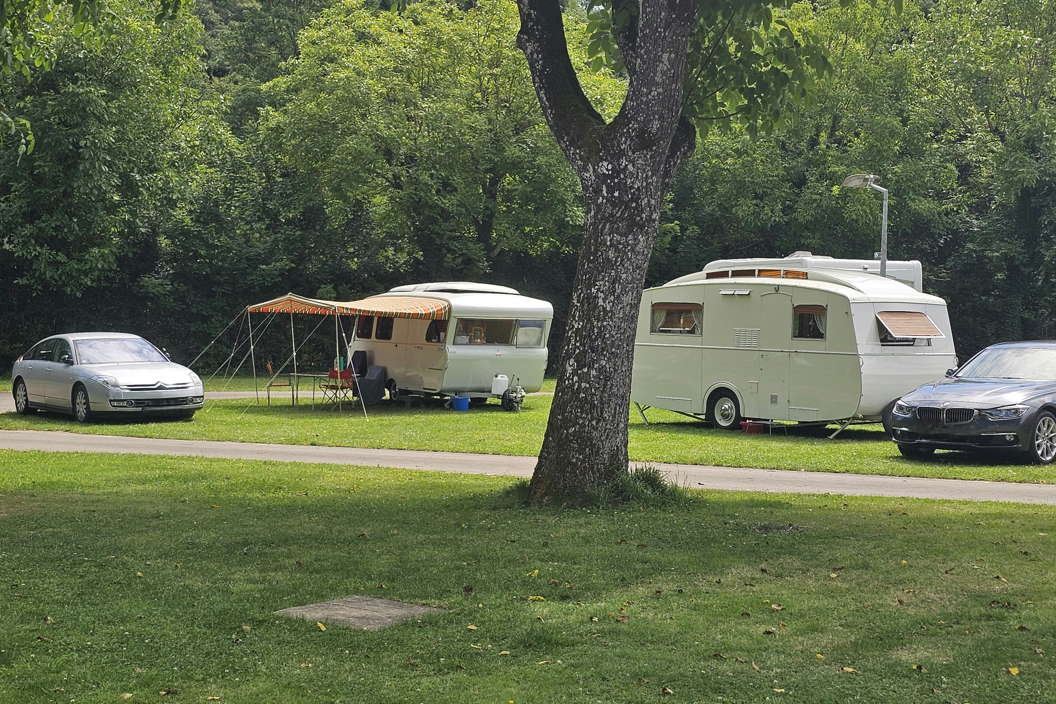 Wohnwagen und Zelt auf einem Campingplatz im Grünen, daneben Autos und ein Weg am Rasen.