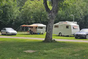 Wohnwagen und Zelt auf einem Campingplatz im Grünen, daneben Autos und ein Weg am Rasen.