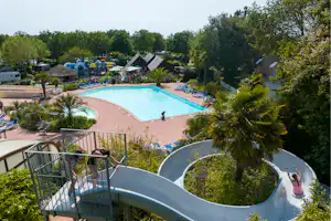 Großer Camping-Pool mit Wasserrutsche, Liegen und Blick auf Bäume, Wohnwagen und Wege im Gartenbereich.
