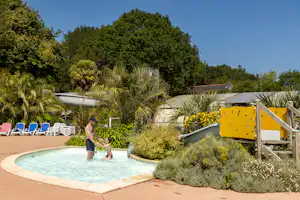 Schwimmbecken am Campingplatz mit Liegen, Palmen und bunten Blumenbeeten, daneben ein Holzsteg und ein Geländer.